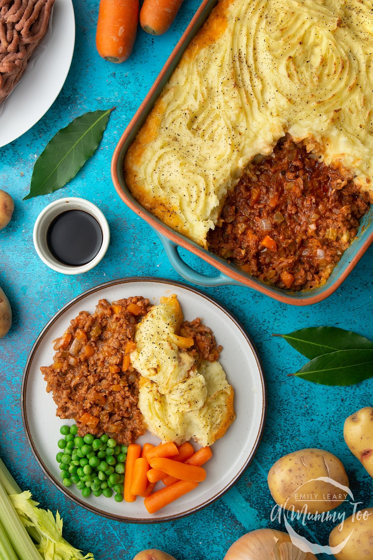 Vegan cottage pie served on a plate with peas and carrots. More pie is shown in its dish. Ingredients are scattered around.