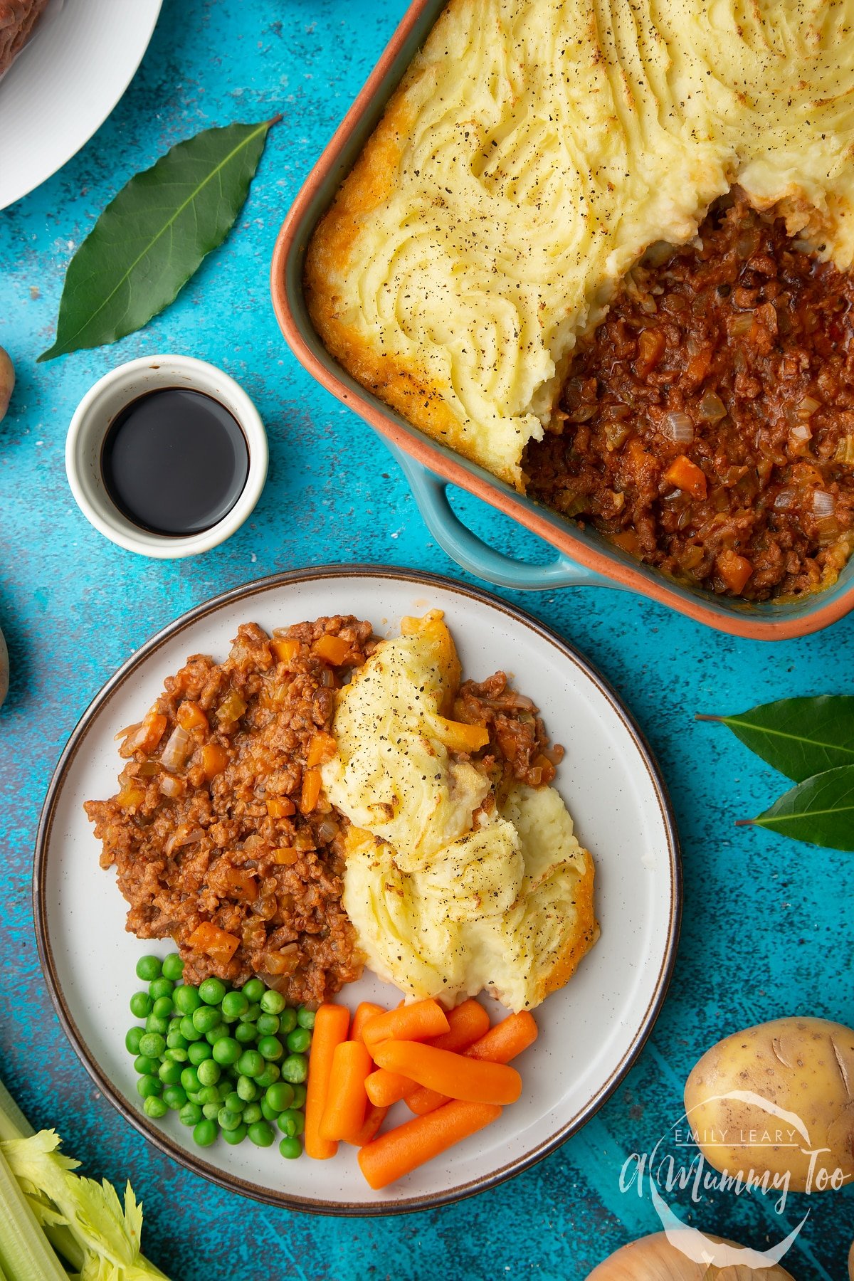 Vegan cottage pie served on a plate with peas and carrots. More pie is shown in a green roasting dish with an orange rim.