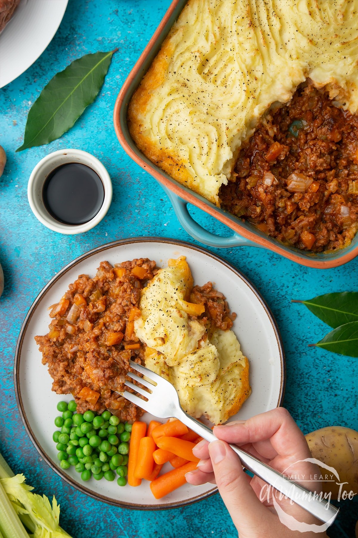 Vegan cottage pie served on a plate with peas and carrots. A hand holding a fork reaches in to take some. More pie is shown in its dish. Ingredients are scattered around.