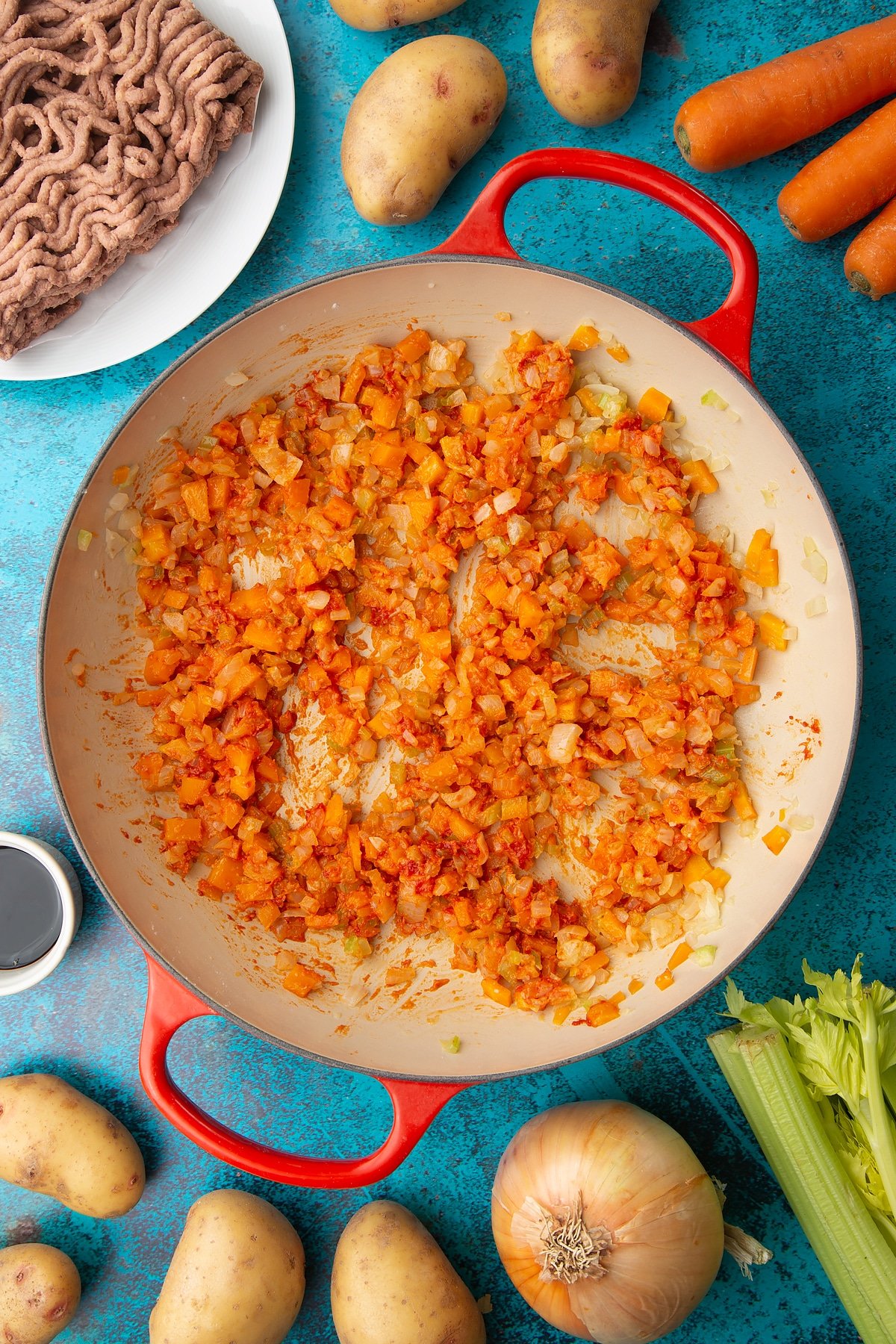 Fried onions, celery, carrots and garlic, flour and tomato puree in a large pan. Ingredients to make vegan cottage pie surround the pan.
