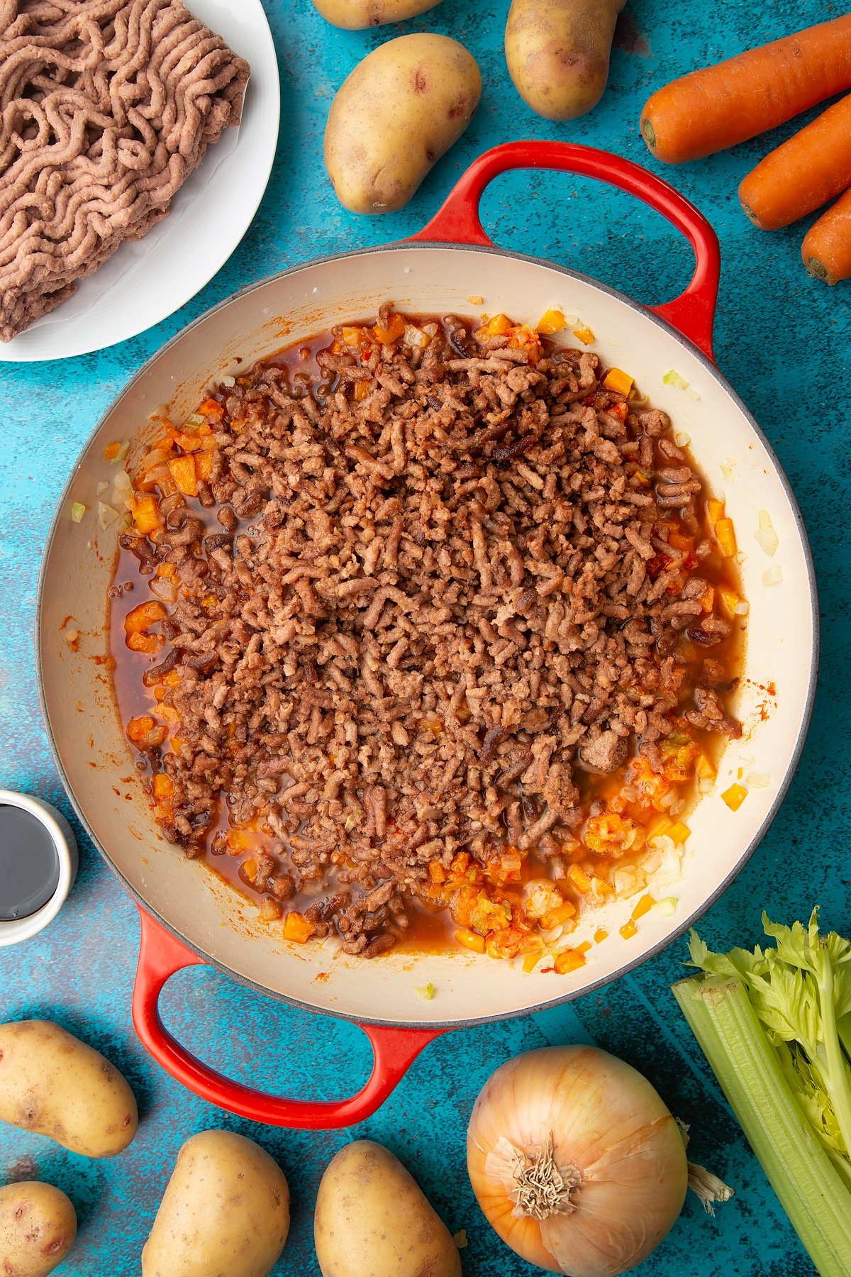 Fried onions, celery, carrots and garlic, flour, tomato puree, meat free mince, sage, wine and stock in a large pan. Ingredients to make vegan cottage pie surround the pan.