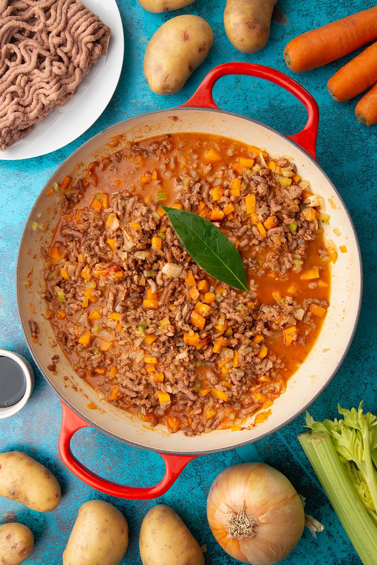 Fried onions, celery, carrots and garlic, flour, tomato puree, meat free mince, sage, wine, soy, bay leaef and stock in a large pan. Ingredients to make vegan cottage pie surround the pan.