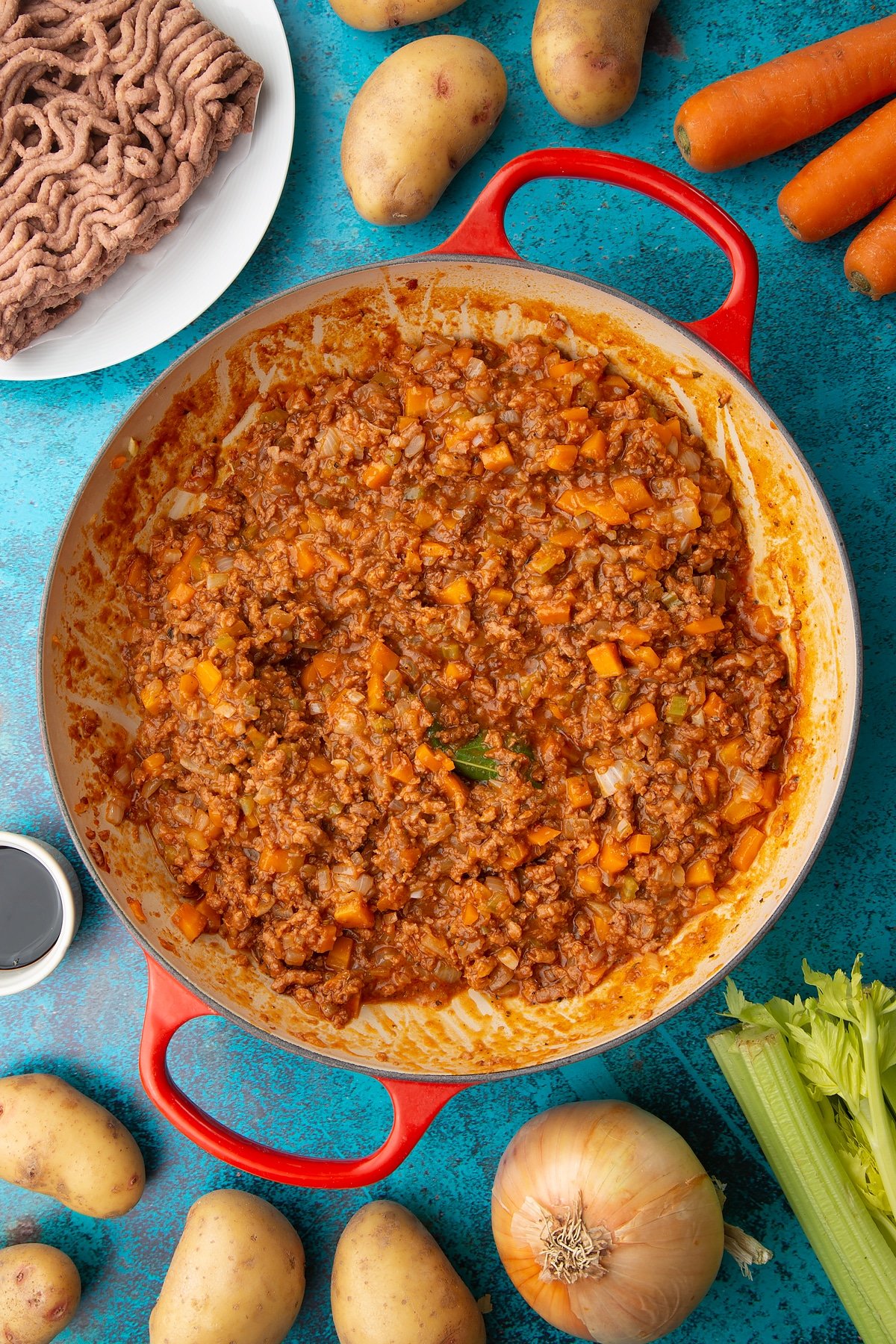 Meat free cottage pie filling in a large pan. Ingredients to make vegan cottage pie surround the pan.