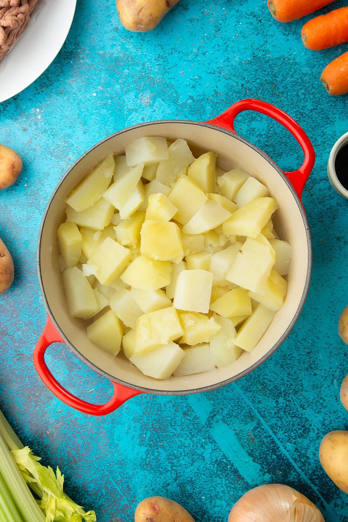 Cooked chopped potatoes in a saucepan. Ingredients to make vegan cottage pie surround the pan.