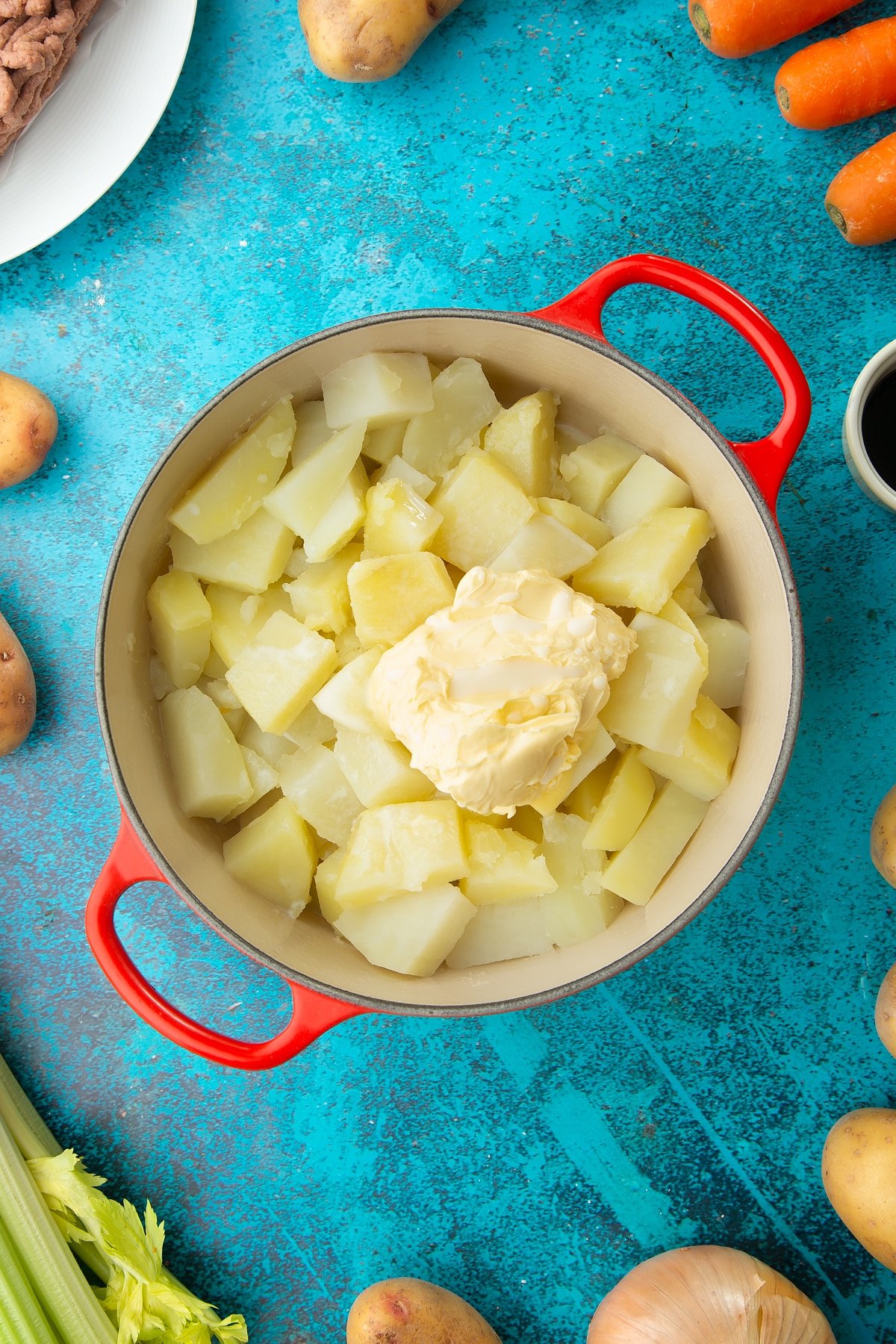 Cooked chopped potatoes, vegan margarine and plant milk in a saucepan. Ingredients to make vegan cottage pie surround the pan.