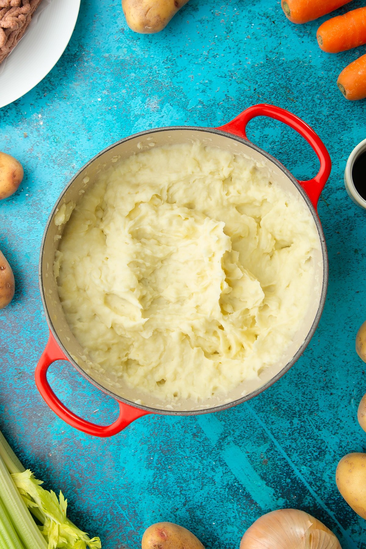 Mashed potatoes in a saucepan. Ingredients to make vegan cottage pie surround the pan.