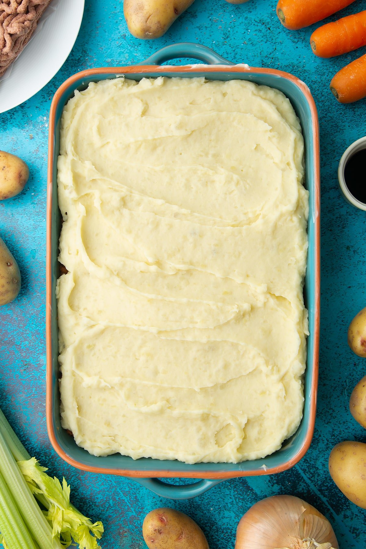 Meat free cottage pie filling topped with mashed potato in a roasting dish. Ingredients to make vegan cottage pie surround the dish.