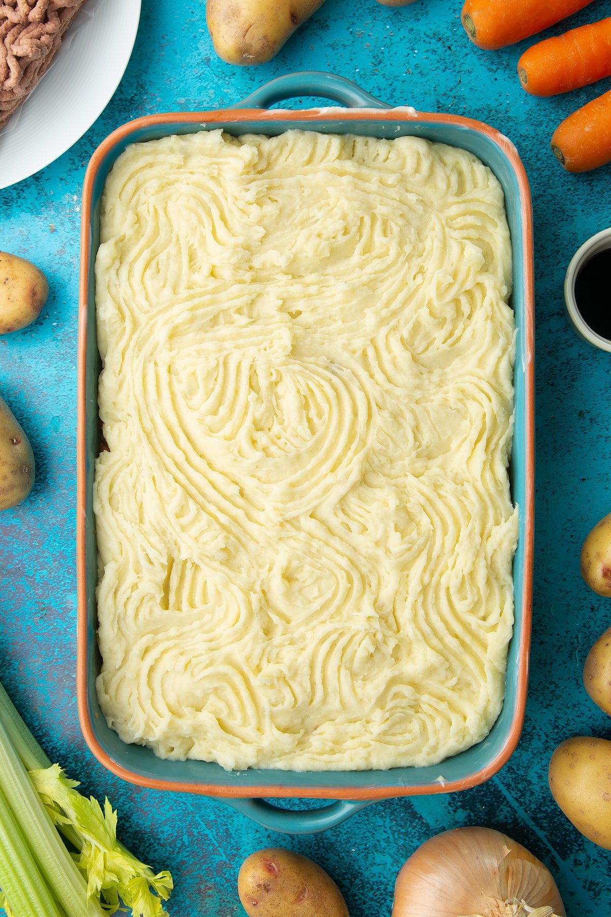 Meat free cottage pie filling topped with mashed potato in a roasting dish. The potato has been textured with a fork. Ingredients to make vegan cottage pie surround the dish.