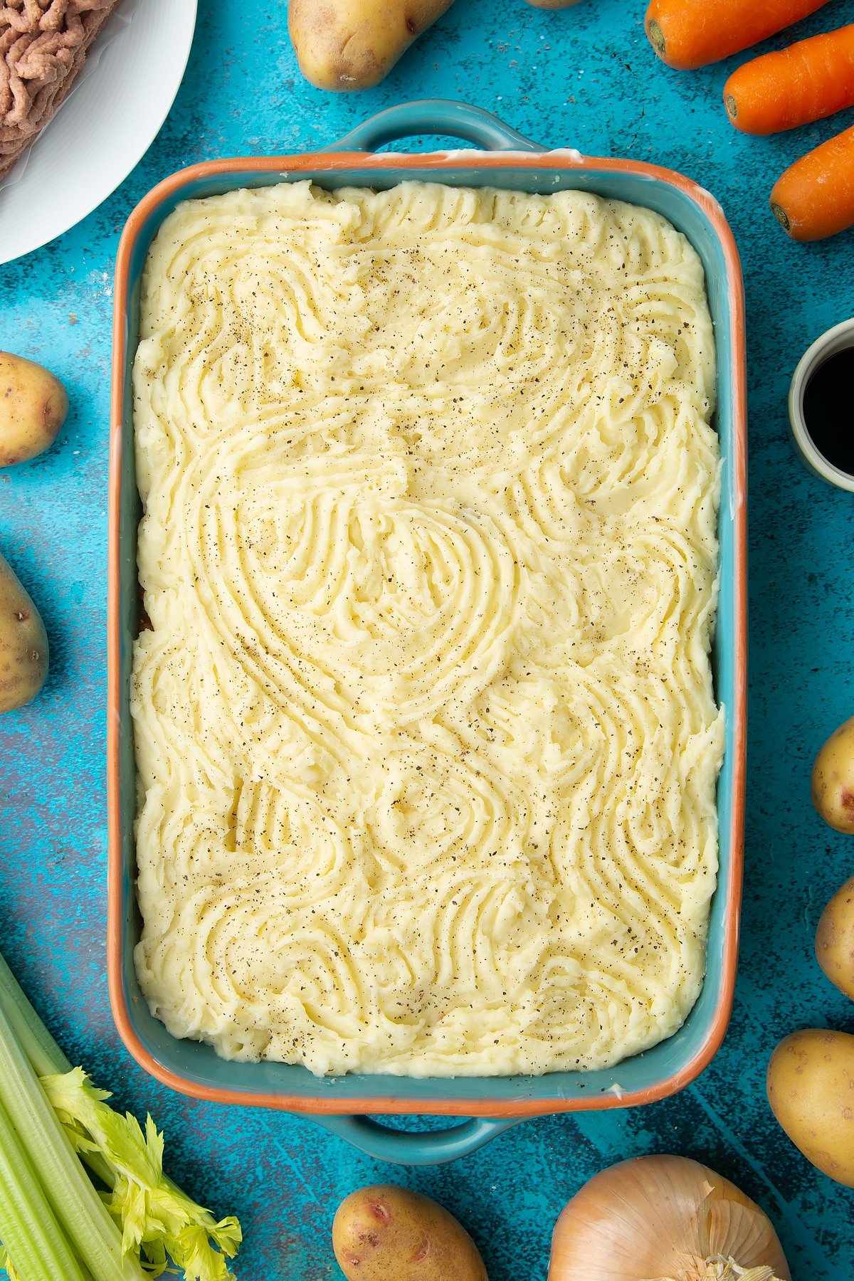 Meat free cottage pie filling topped with mashed potato in a roasting dish. The potato has been textured with a fork and seasoned with pepper. Ingredients to make vegan cottage pie surround the dish.