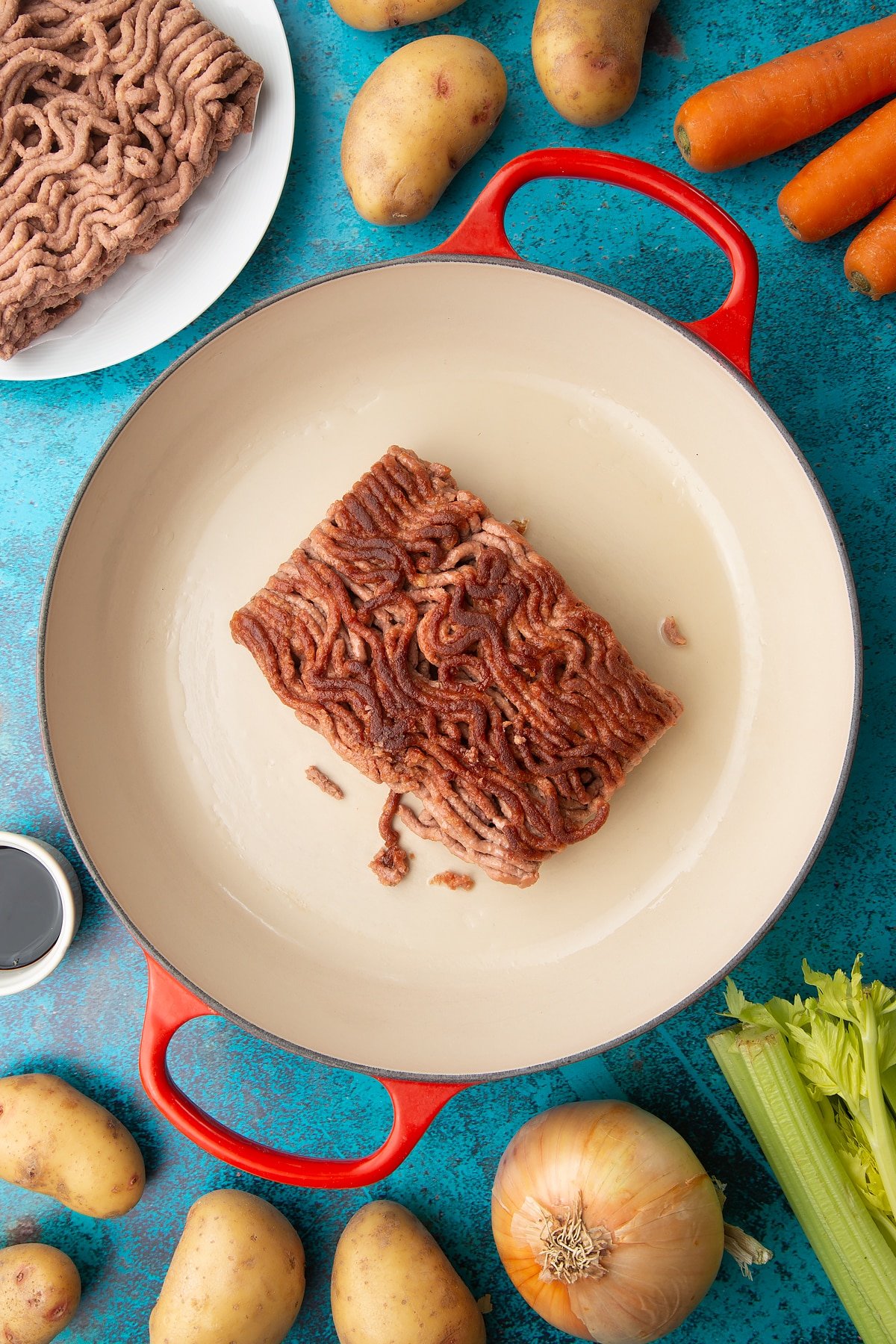 A block of browned meat free mince in a large pan. Ingredients to make vegan cottage pie surround the pan.