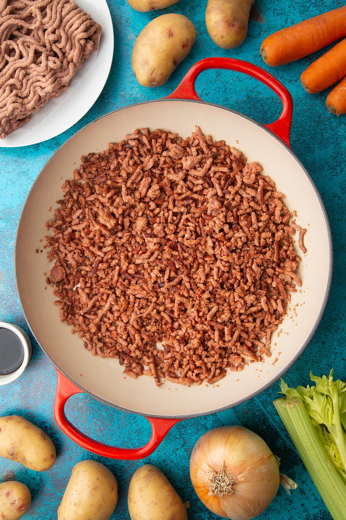 Crisp and browed meat free mince in a large pan. Ingredients to make vegan cottage pie surround the pan.