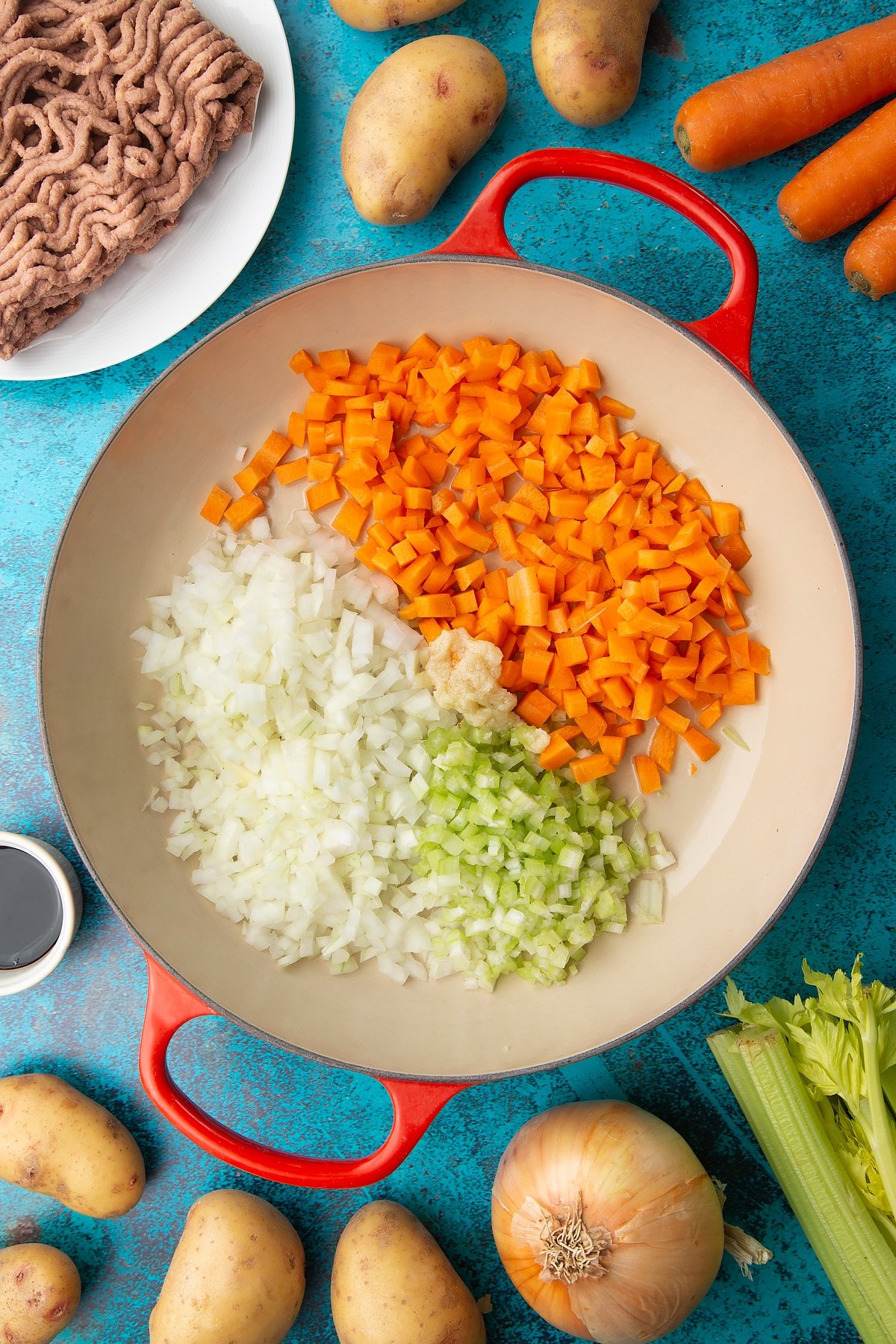 Chopped onions, celery, carrots and garlic in a large pan. Ingredients to make vegan cottage pie surround the pan.