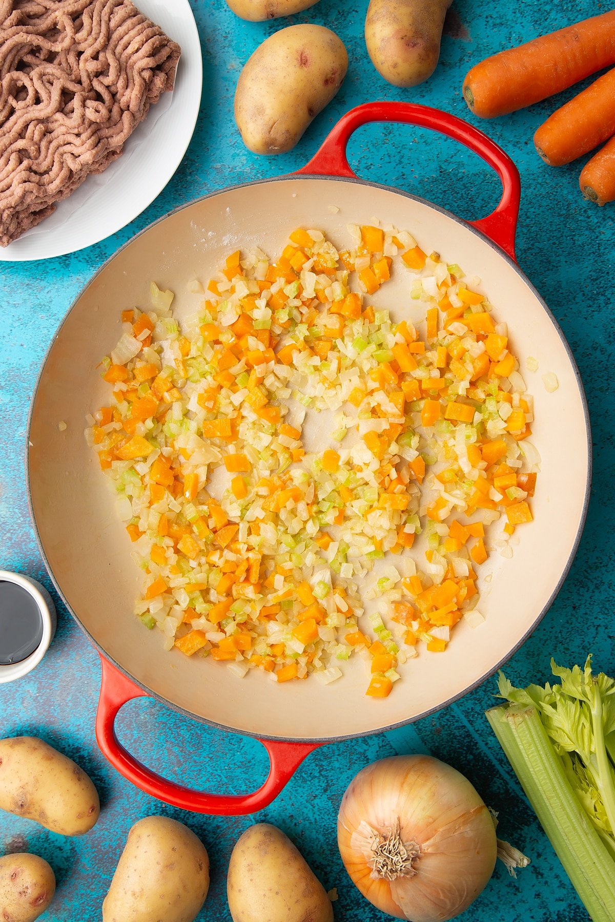 Fried onions, celery, carrots and garlic in a large pan. Ingredients to make vegan cottage pie surround the pan.