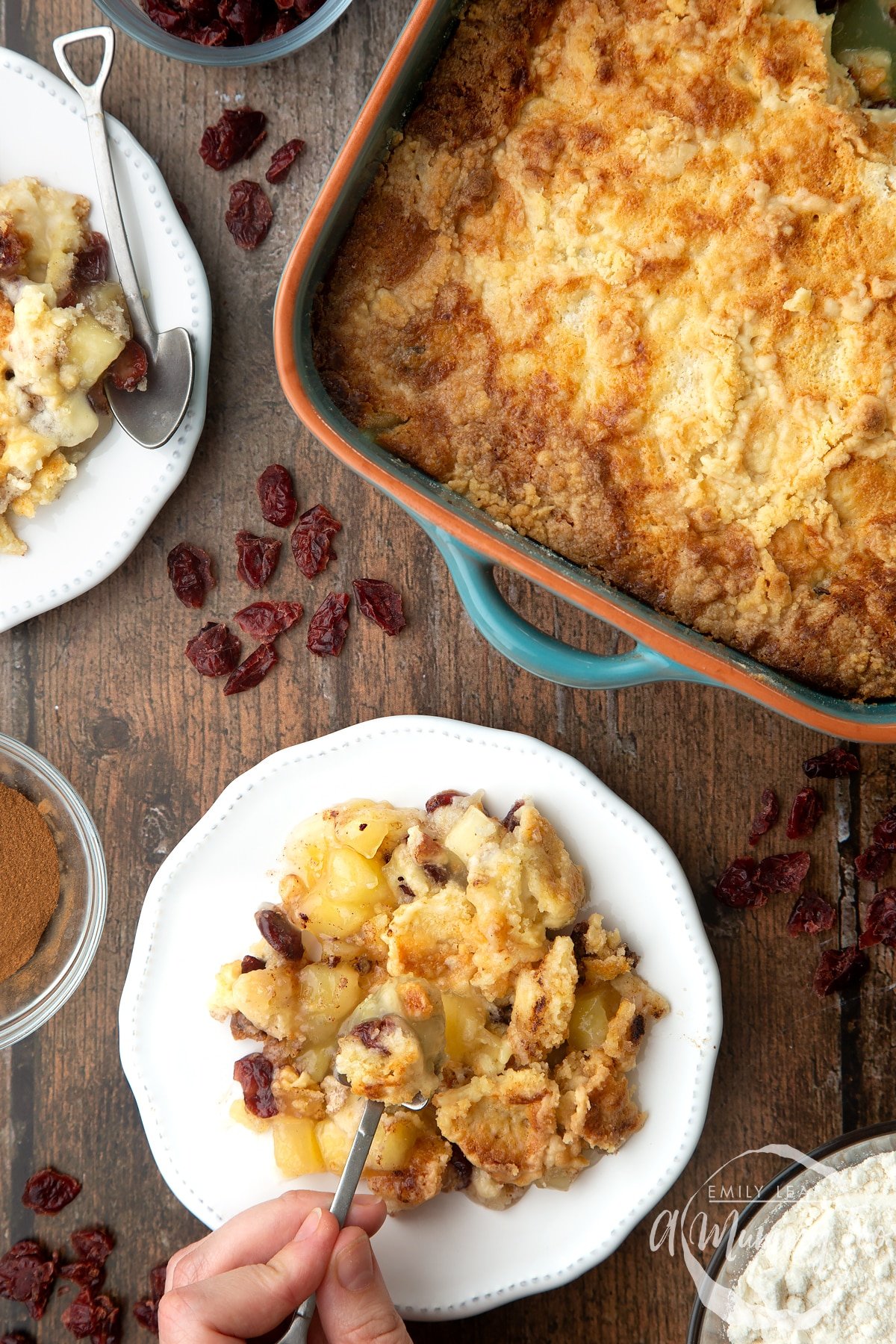 Apple cranberry dump cake served onto a small white plate. A hand holds a small spoon, which is delving into the cake. A tray of the cake sits to the side.