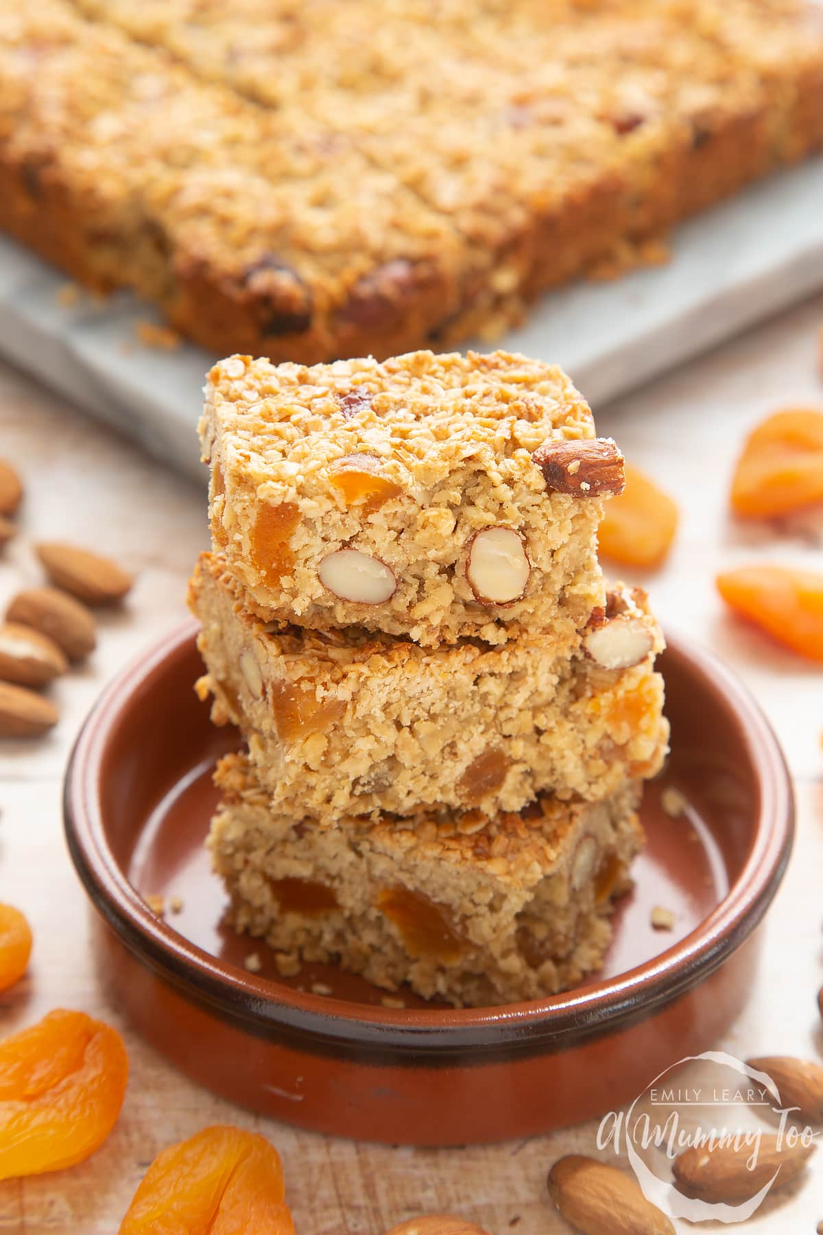 Front angle shot of Apricot oat slices stacked in a brown bowl with a mummy too logo in the lower-right corner