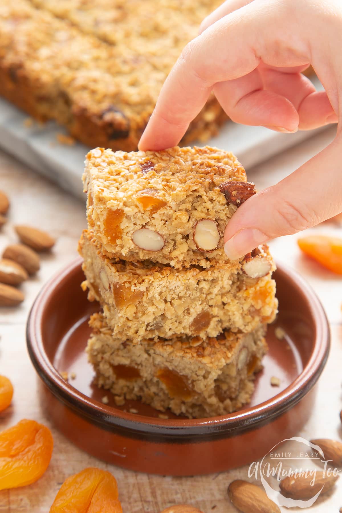 Front angle shot of a hand holding an Apricot oat slice served on a brown bowl with a mummy too logo in the lower-right corner