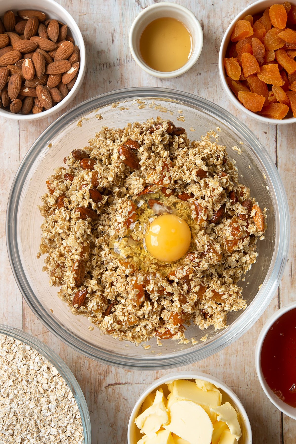 Overhead shot of oats, liquid mixture, apricots, nuts, and egg in a large clear bowl