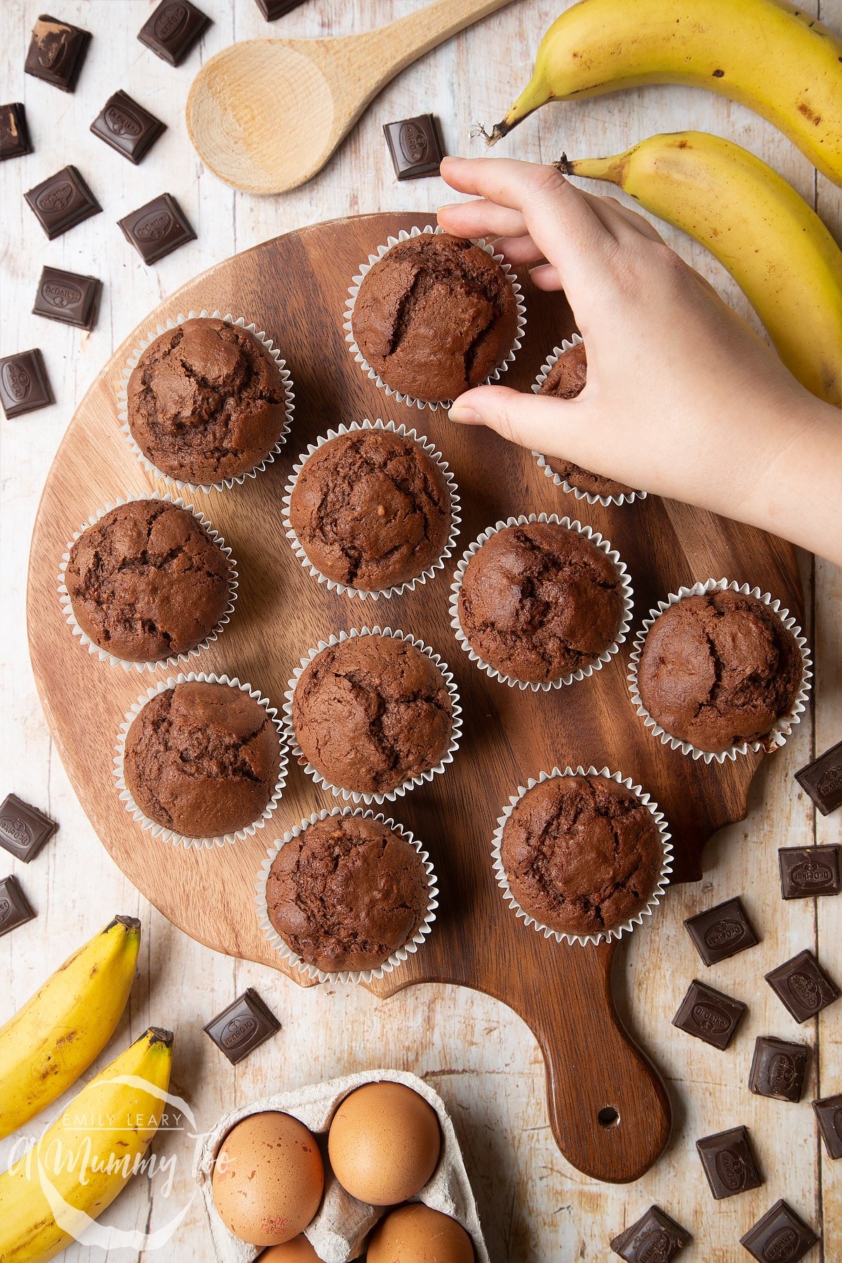 Overhead shot of a hand holding Banana and chocolate muffins served on a wooden plate with a mummy too logo in the lower-right corner