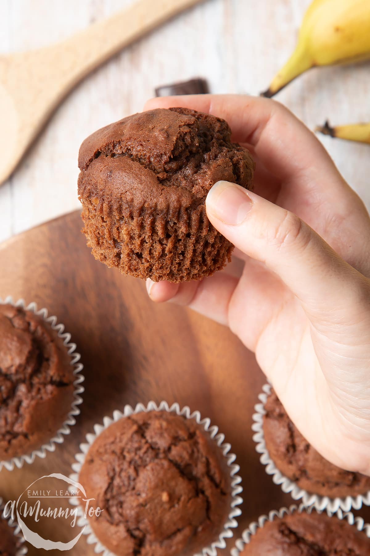 Overhead shot of a hand holding a Banana and chocolate muffin served on a wooden plate with a mummy too logo in the lower-left corner