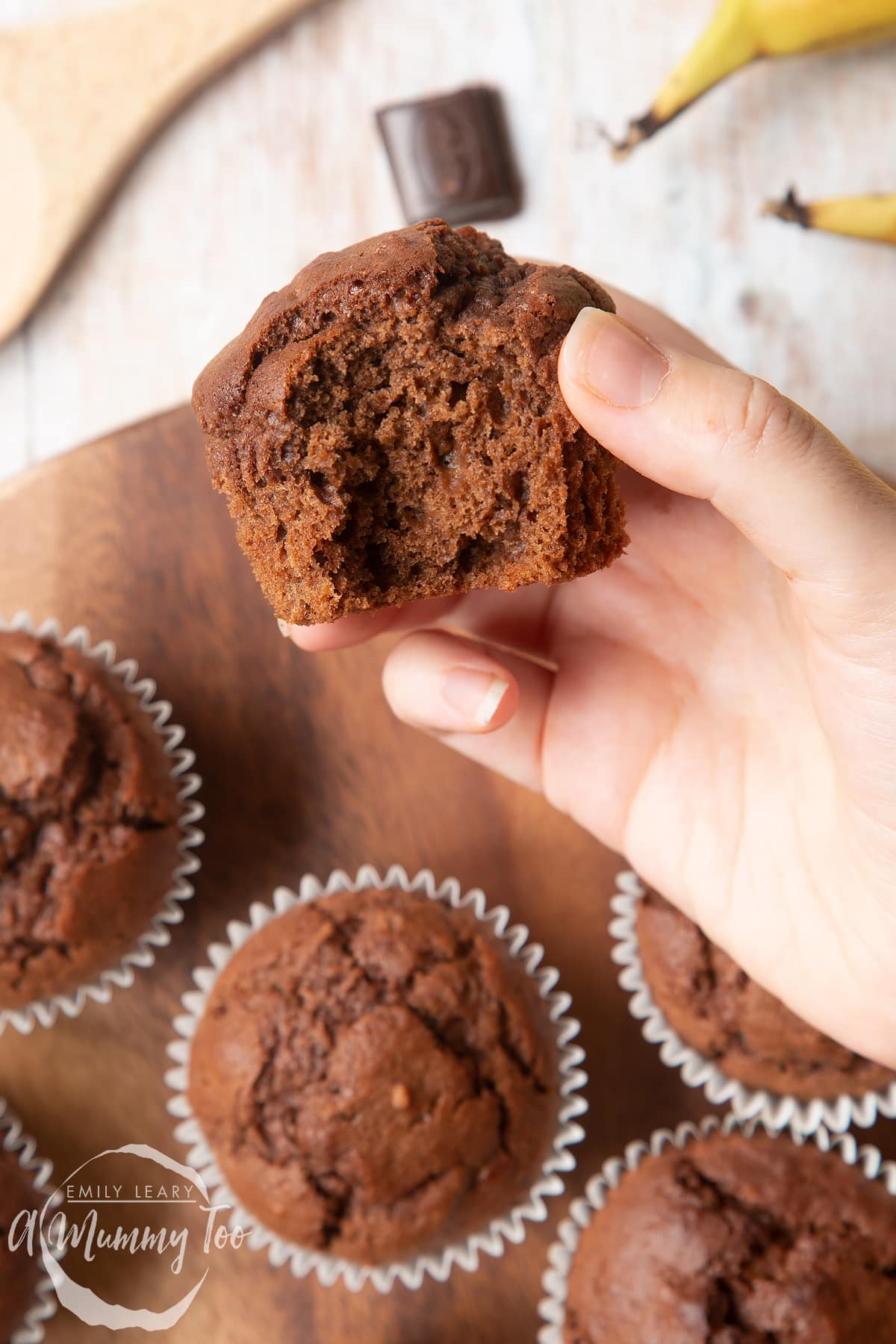 front angle shot of a hand holding a half eaten banana and chocolate muffins with a mummy too logo in the lower-left corner