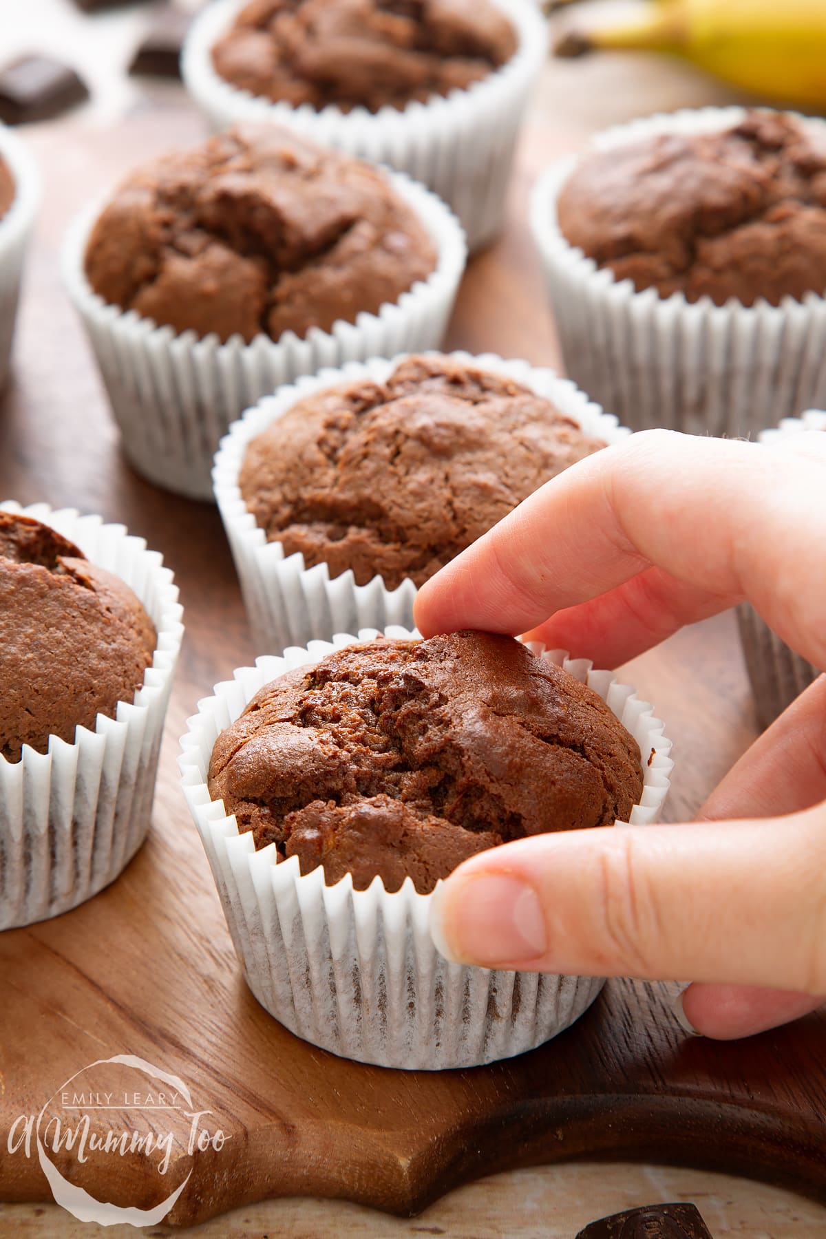 Front angle shot of a hand holding Banana and chocolate muffins served on a wooden plate with a mummy too logo in the lower-right corner