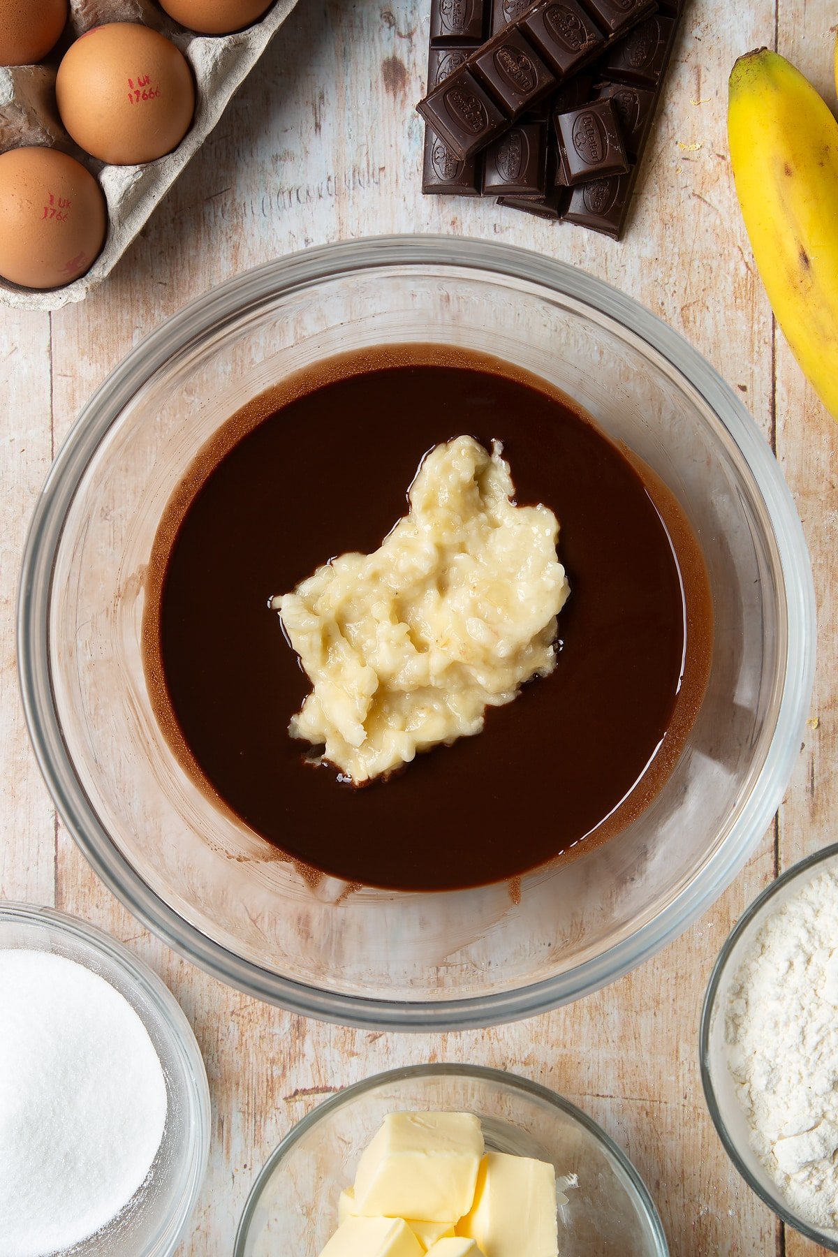 Overhead shot of chocolate mix and mashed bananas in a large clear bowl