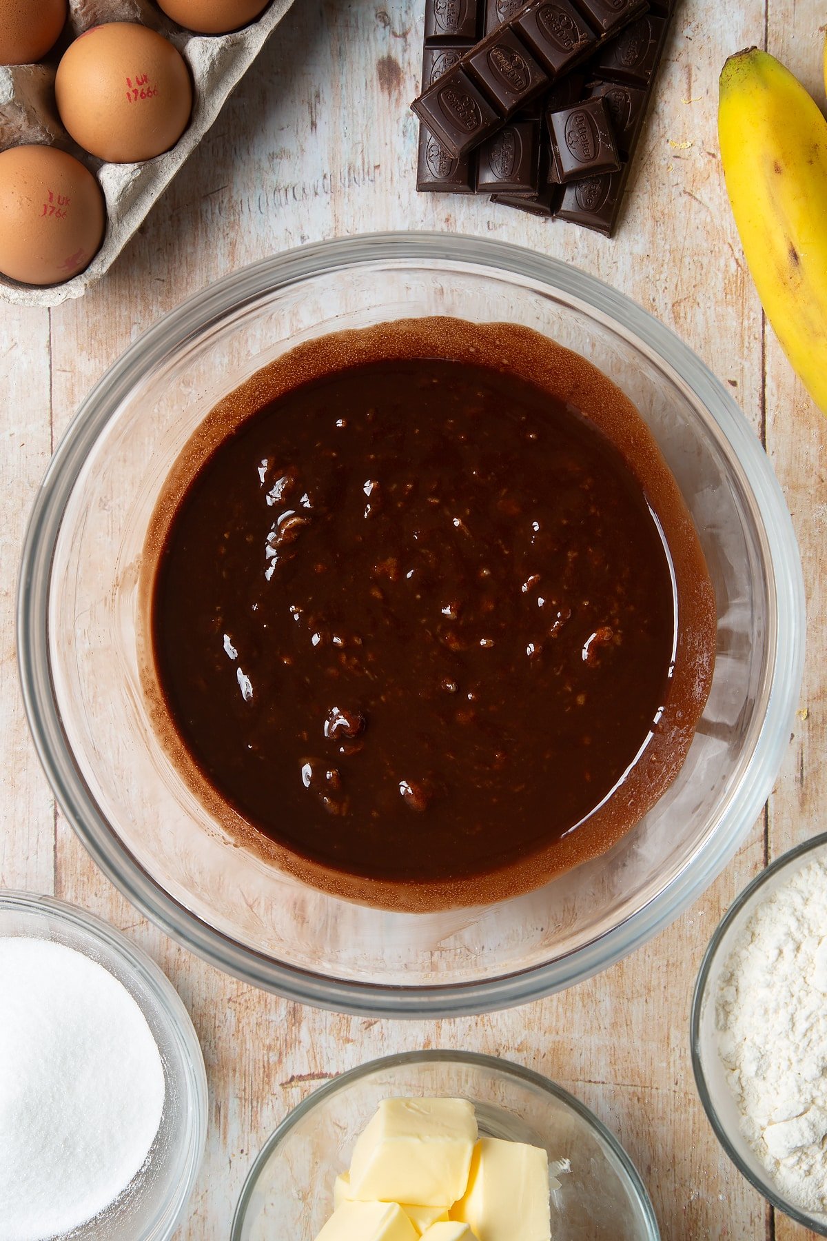 Overhead shot of chocolate liquid mixed with mashed bananas in a large clear bowl