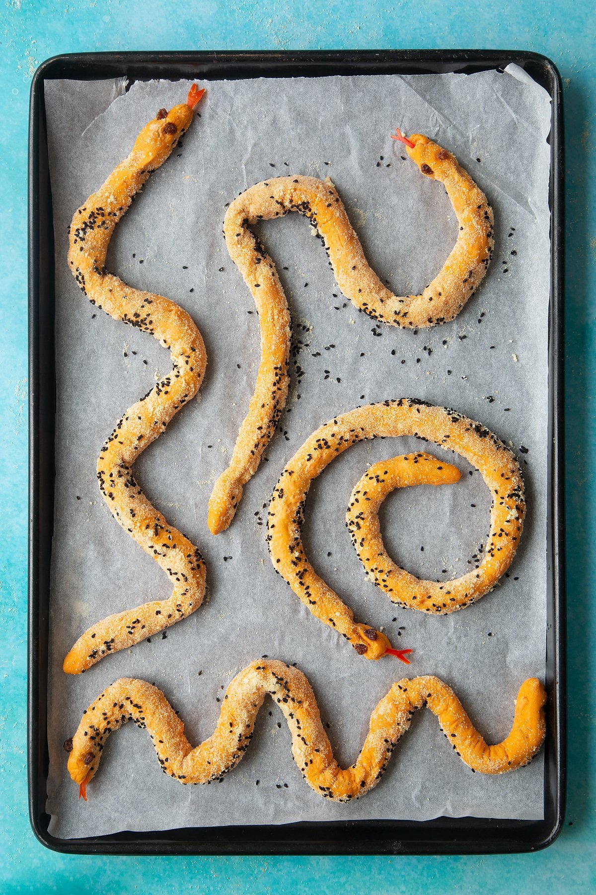 A lined baking tray with paprika and carrot bread dough rolled into a long sausage and decorated to look like snakes. 