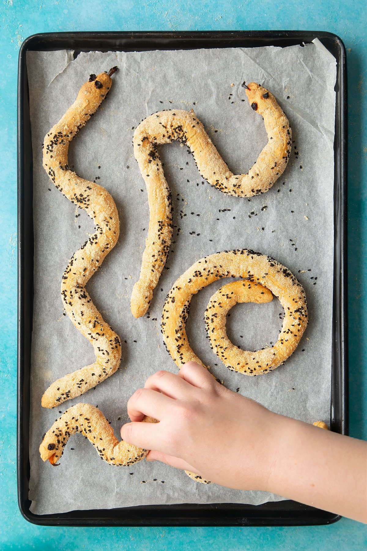 A lined baking tray with freshly baked bread snakes. A hand reaches to take one.