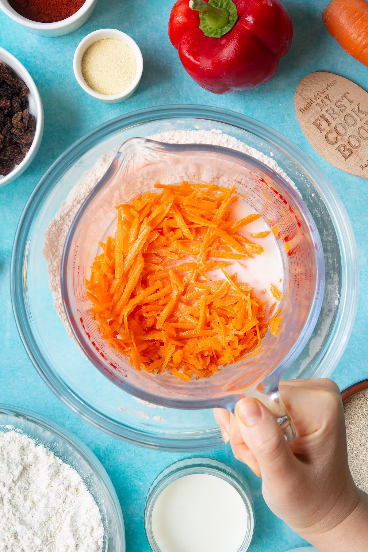 A hand holds a jug containing grated carrot, milk and water. Flour, salt, paprika and yeast are in a mixing bowl below. Ingredients to make bread snakes surround the bowl.