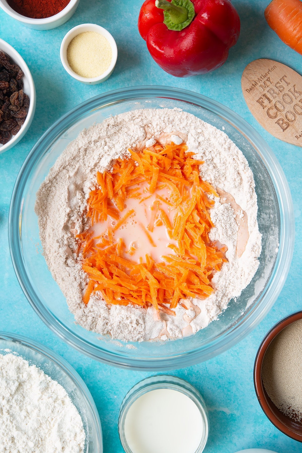 Flour, salt, paprika, yeast, grated carrot, milk and water in a mixing bowl. Ingredients to make bread snakes surround the bowl.