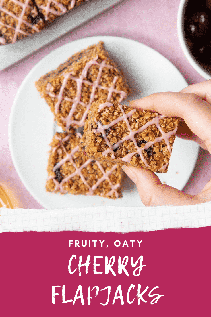 Overhead shot of a hand holding a cherry flapjack with graphic text FRUITY, OATY CHERRY FLAPJACKS below