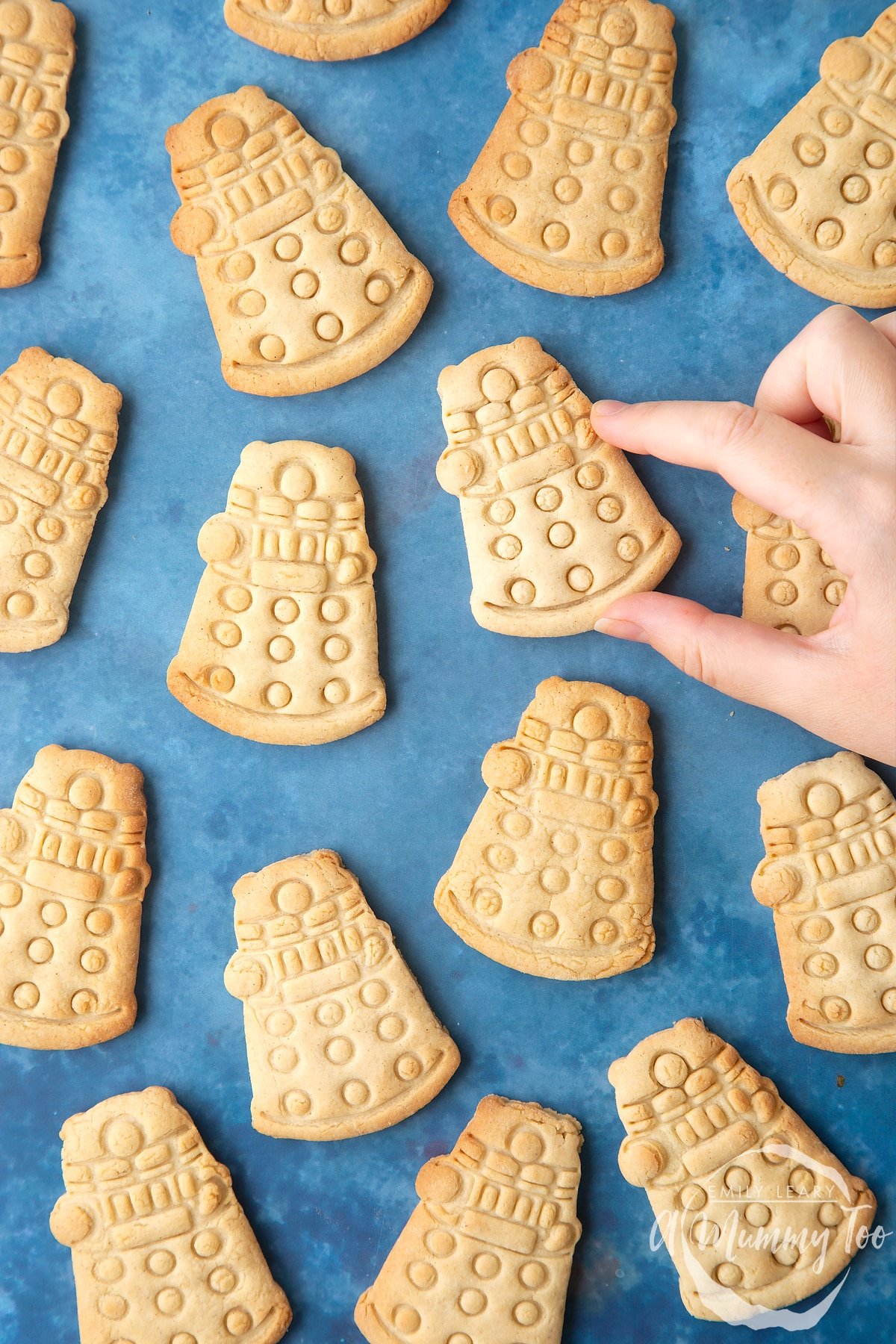 Dalek cookies on a blue background. The cookies are undecorated. A hand reaches to take one.