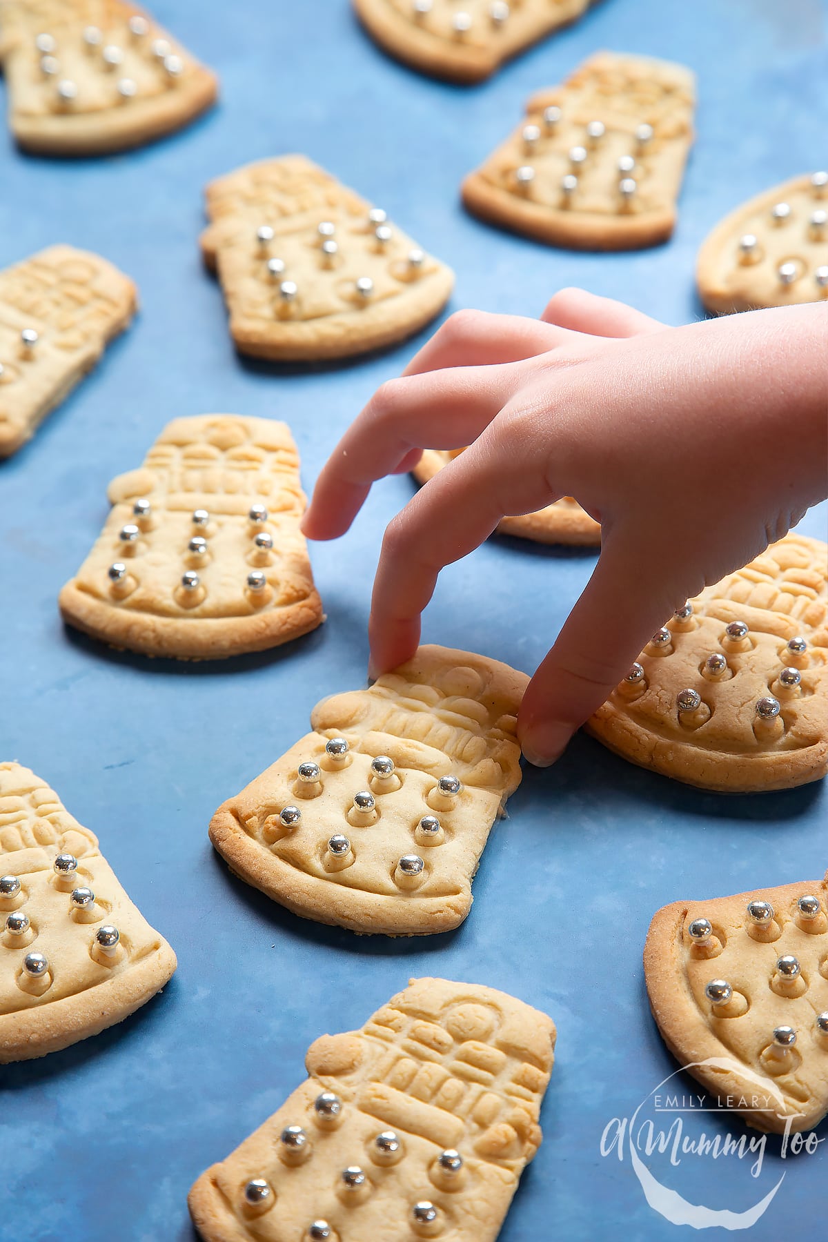 Dalek cookies on a blue background. The cookies are decorated with edible silver candy balls adhered with white icing. A hand reaches to take one.