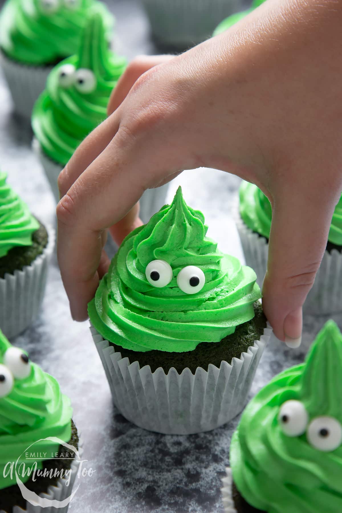 Green monster cakes made with dyed-green chocolate chip cupcakes topped with green peppermint frosting with added candy eyes. A hand reaches to take one.