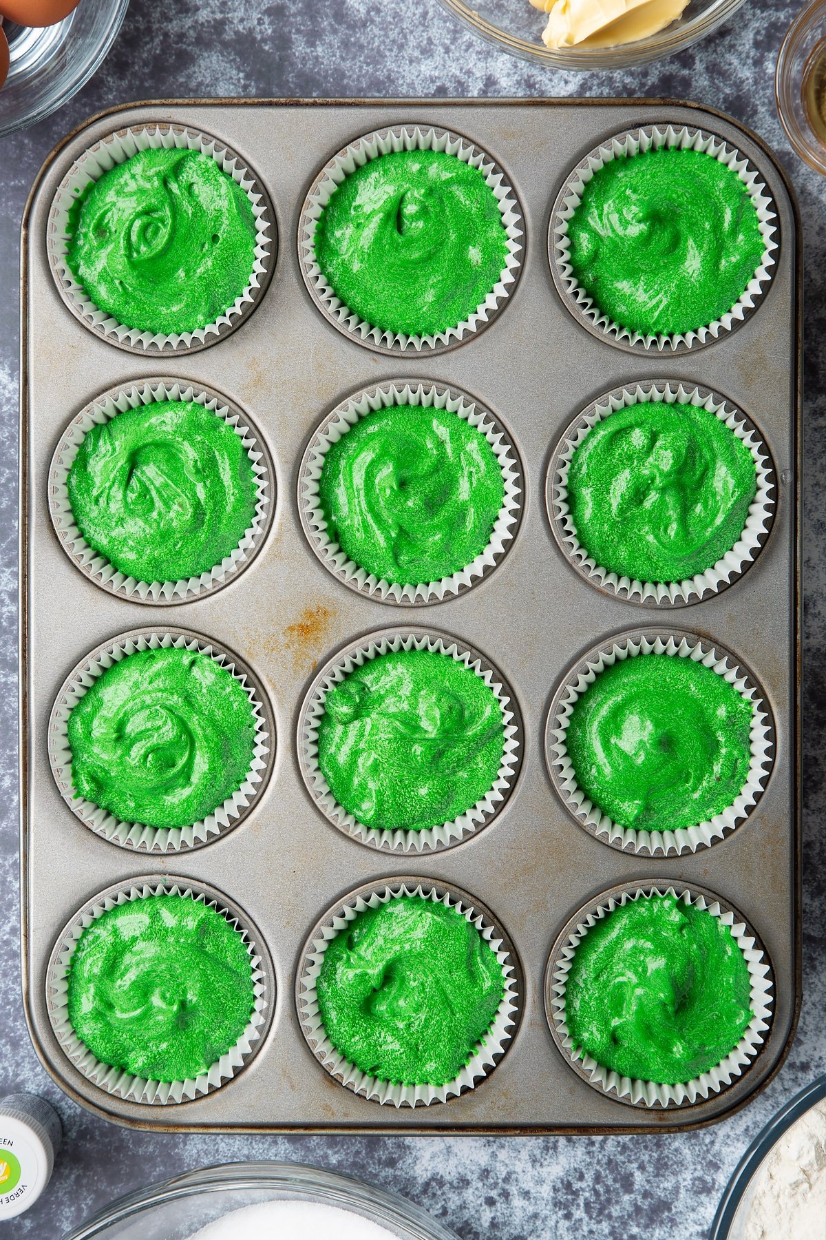 12 hole muffin tray lined with cupcake cases filled with green vanilla chocolate chip cake batter. Ingredients to make green monster cakes surround the tray. 