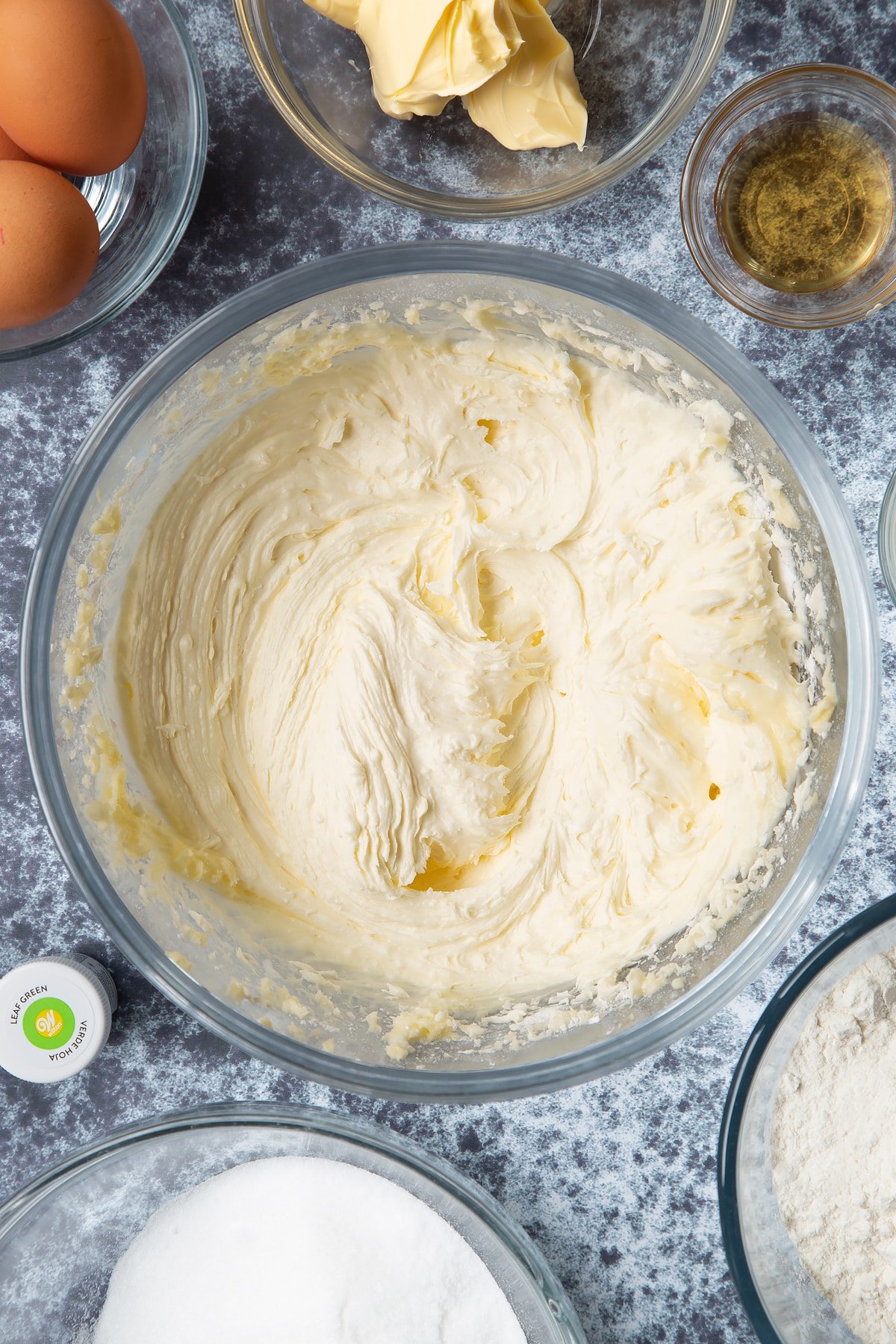 Sugar, butter and peppermint extract whisked together in a large mixing bowl. Ingredients to make green monster cakes surround the bowl. 