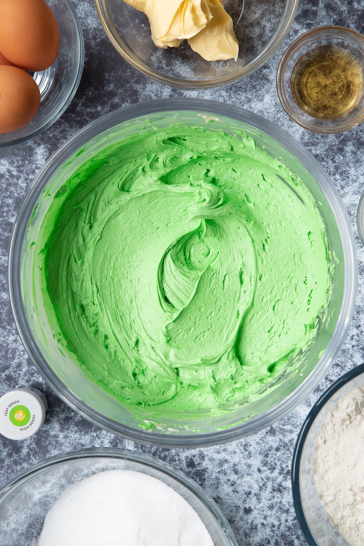 Green peppermint buttercream in a large mixing bowl. Ingredients to make green monster cakes surround the bowl. 