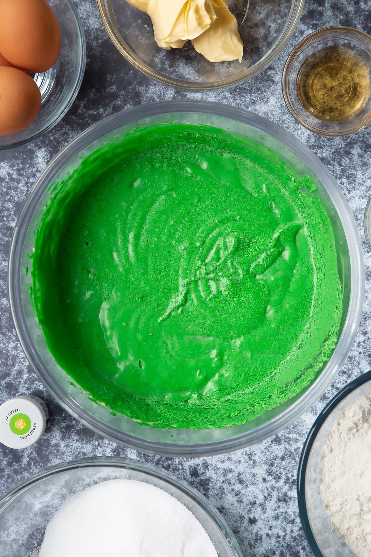 Green dyed vanilla cake batter in progress in a large mixing bowl. Ingredients to make green monster cakes surround the bowl. 