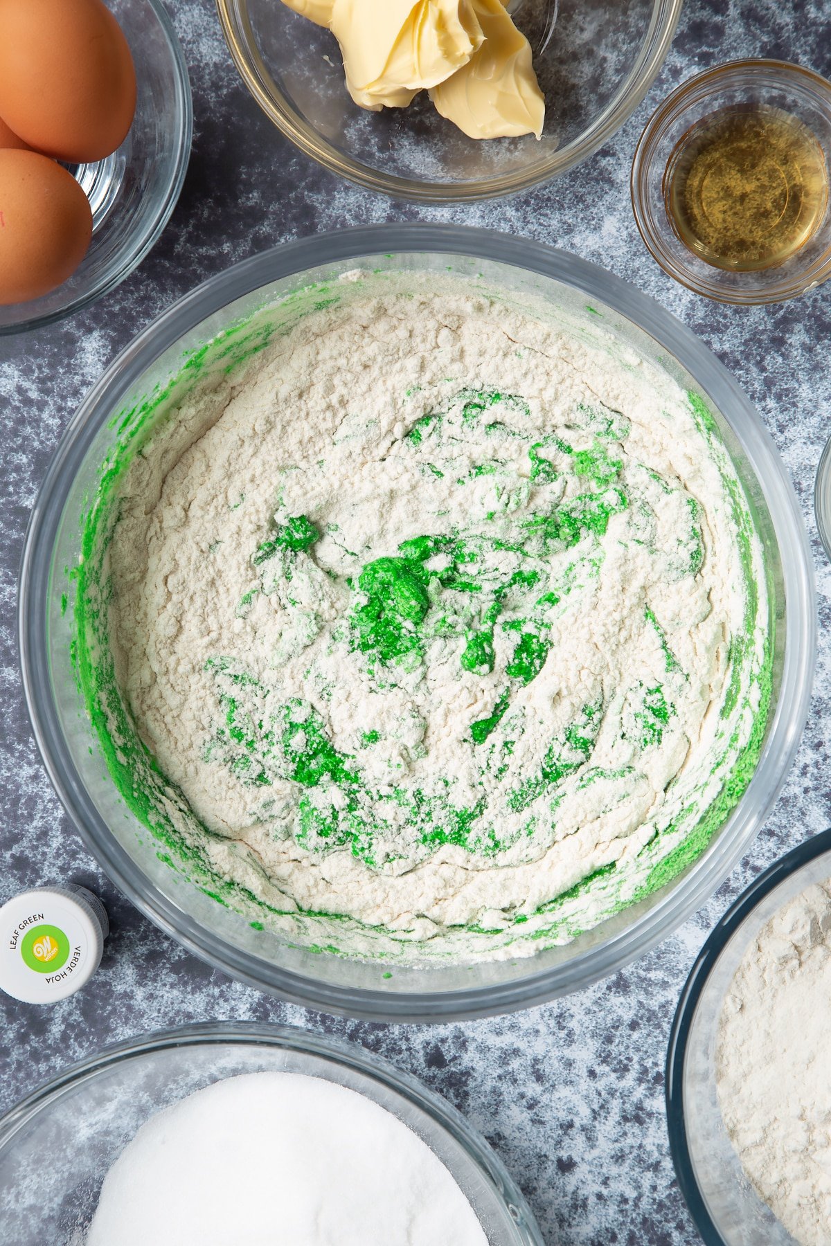 Green dyed vanilla cake batter with flour in progress in a large mixing bowl. Ingredients to make green monster cakes surround the bowl.