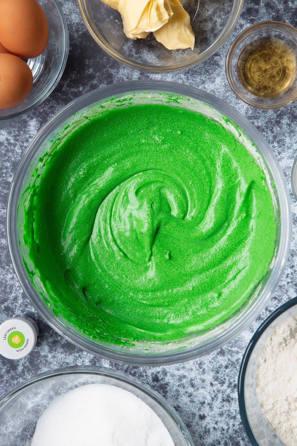 Green dyed vanilla cake batter in a large mixing bowl. Ingredients to make green monster cakes surround the bowl. 