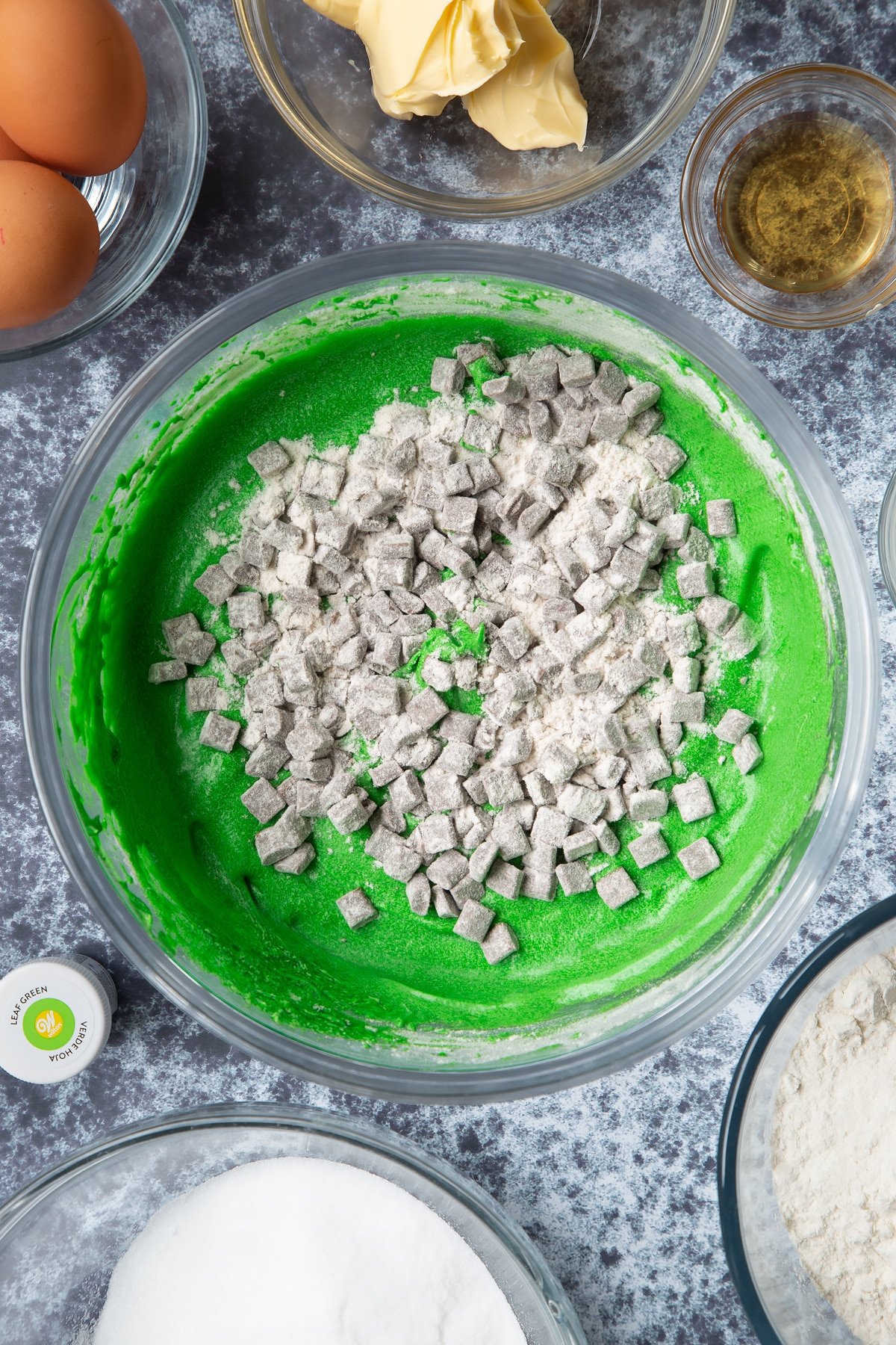 Green dyed vanilla cake batter with floured chocolate chips on top in a large mixing bowl. Ingredients to make green monster cakes surround the bowl. 