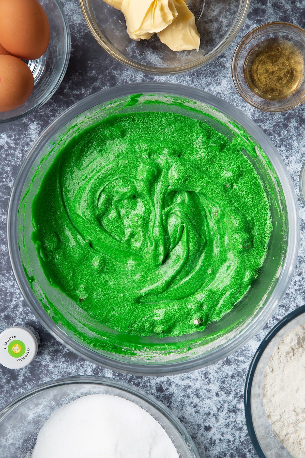 Green dyed vanilla chocolate chip cake batter in a large mixing bowl. Ingredients to make green monster cakes surround the bowl. 