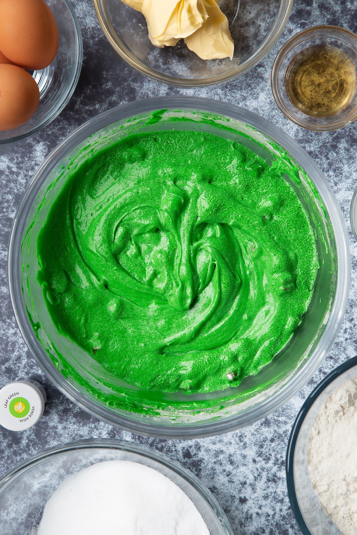 Green dyed vanilla chocolate chip cake batter in a large mixing bowl. Ingredients to make green monster cakes surround the bowl.
