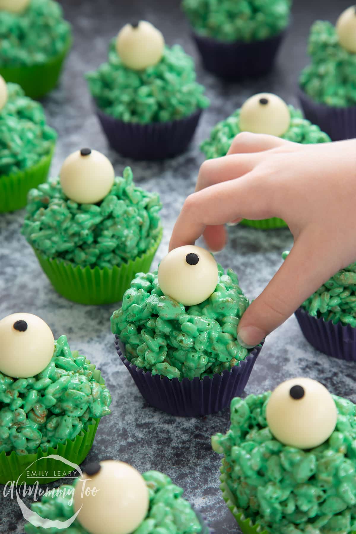 Halloween crispy cakes in purple and green cupcake cases. The crispy cakes are dyed green and topped with white chocolate spheres decorated to look like eyeballs. A little hand reaches for one.