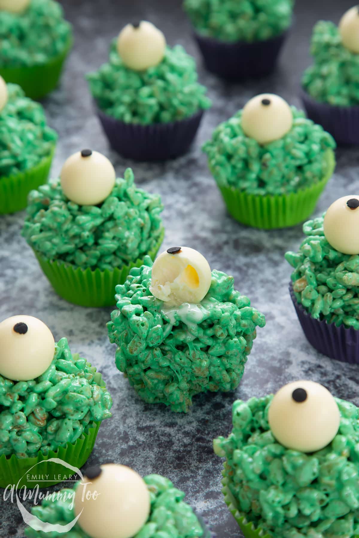 Halloween crispy cakes in purple and green cupcake cases. The crispy cakes are dyed green and topped with white chocolate spheres decorated to look like eyeballs. One has been bitten.