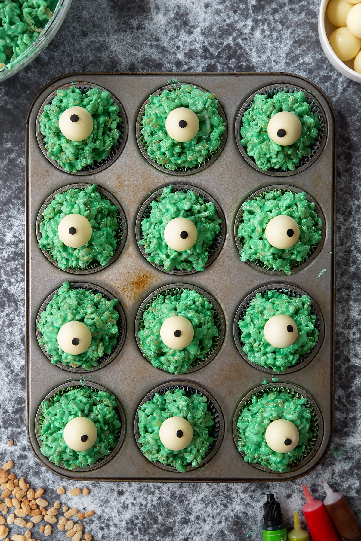 12-hole muffin tray lined with cupcake cases filled with green rice crispy treats topped with white chocolate spheres decorated with icing pupils. Ingredients to make Halloween crispy cakes surround the tray.