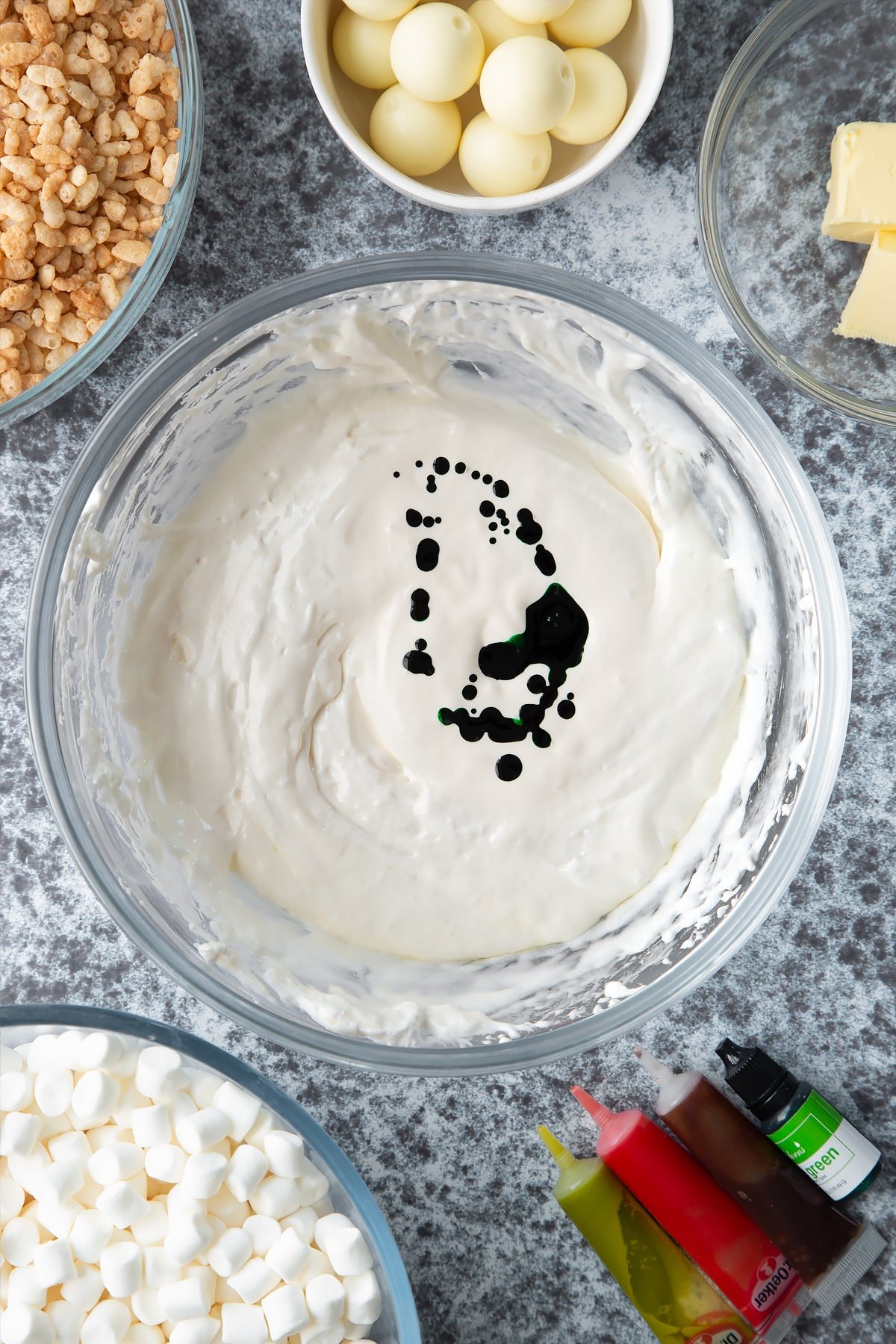 Melted marshmallows and green food colouring in a large mixing bowl. Ingredients to make Halloween crispy cakes surround the bowl.