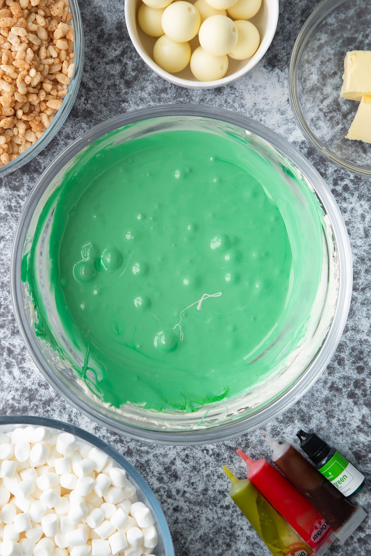 Melted marshmallows dyed green in a large mixing bowl. Ingredients to make Halloween crispy cakes surround the bowl.