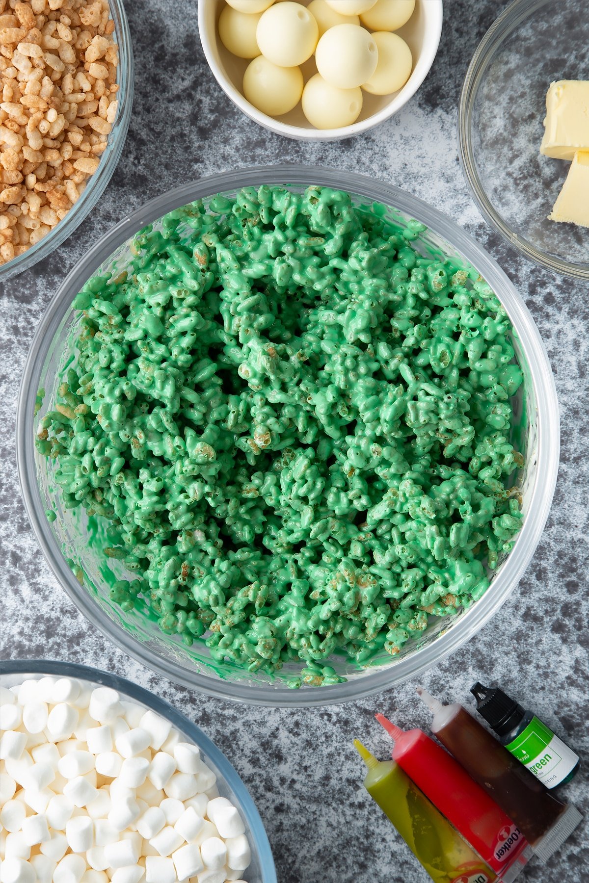 Dyed-green rice crispie treat mix in a large mixing bowl. Ingredients to make Halloween crispy cakes surround the bowl.